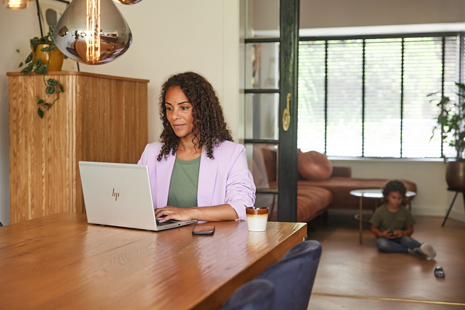 Vrouw zittend aan tafel achter haar laptop met haar telefoon en kop koffie naast zich