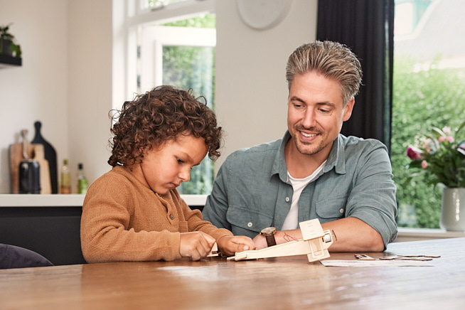 Man met zoon zittend aan tafel en beide kijkend naar een speelgoedvliegtuig