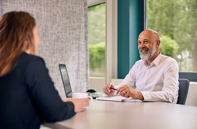 Mevrouw in gesprek met een vriendelijk ogende financieel adviseur. De adviseur zit achter een laptop en heeft een notitieblok bij zich.