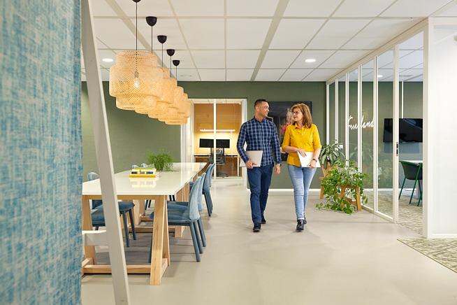 Man en vrouw die samen door een kantoorpand aan het wandelen zijn. Ze zijn met elkaar in gesprek en houden beiden hun laptop in de hand