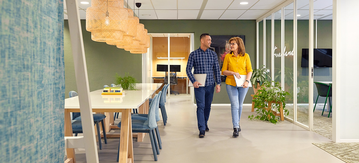 Twee collega's wandelend door kantoor terwijl ze een gesprek voeren met hun laptop in hun handen