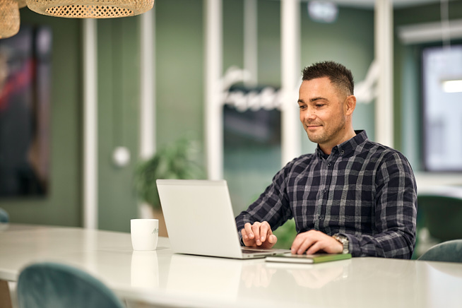 Meneer die glimlachend aan een tafel achter zijn laptop aan het werk is