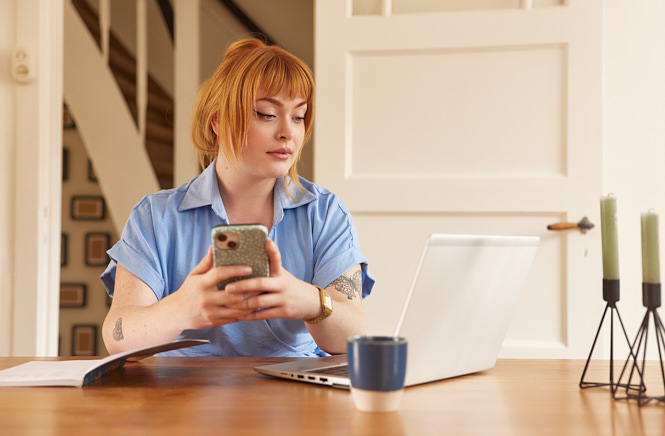 Vrouw zittend aan tafel met mobiel in hand kijkend naar haar laptop