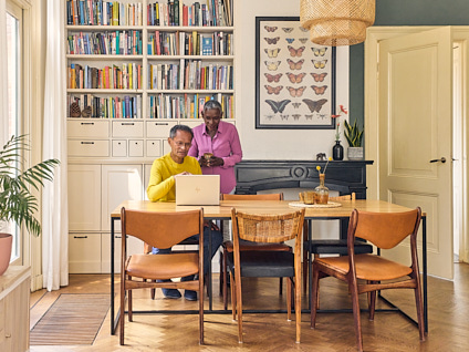 Ouder koppel waarvan een staand en een zittend aan tafel achter hun laptop