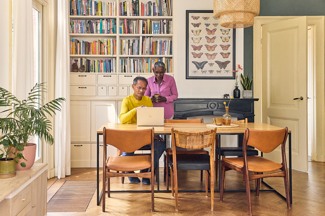 Zittende man en staande vrouw kijken samen naar een opengeklapt laptopscherm in hun woonkamer