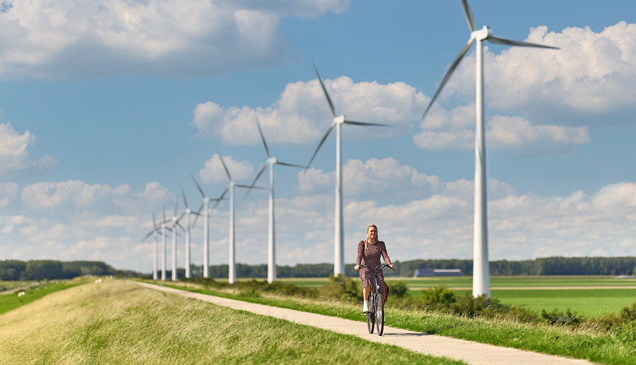 Vrouw op een fiets die over een fietspad fietst op een dijk met windmolens achter zich