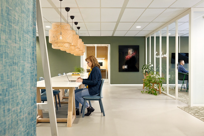 Mevrouw die aan het werk is op haar laptop aan een lange tafel. Op de achtergrond zit een meneer in een vergaderruimte