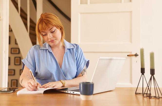 Vrouw zittend aan tafel schrijvend in een notitieblok met een laptop naast zich