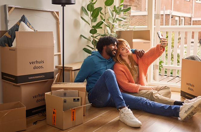 Man en vrouw zittend op de grond tussen de verhuisdozen terwijl ze een selfie maken
