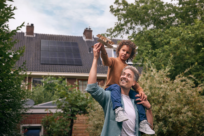 Man met zoon op schouders die samen een speelgoedvliegtuig vasthouden en besturen voor een woning met zonnepanelen